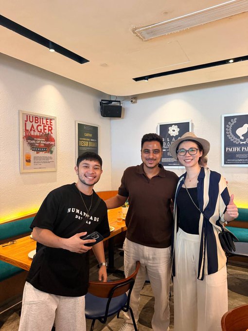Photo of three people smiling in front of a wall of vintage beer advertisements, with the leftmost individual wearing a 'Human Made' t-shirt.