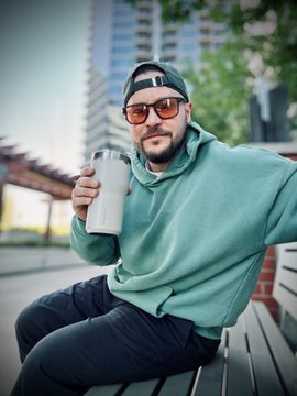 A man in a green hoodie, backward cap, and sunglasses sits on a bench holding a white tumbler, channeling a modern cowboy vibe in an urban setting—capturing the "Stampede" spirit and Back to the Future Part III-inspired fit mentioned in the post, while sipping his coffee amid the Friday grind.