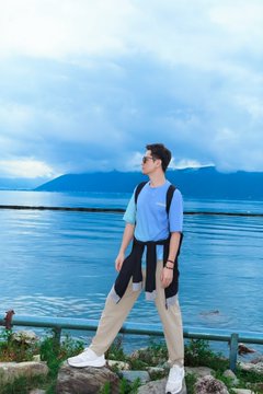 A man wearing sunglasses, a light blue t-shirt, and beige pants stands by a railing overlooking a large blue lake with mountains in the distance.