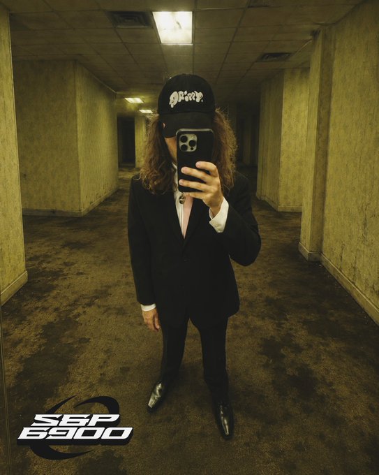 A mirror selfie of a person in a black suit and cap taking a photo with their phone in a dimly lit parking garage, with "SBP 6900" text at the bottom.