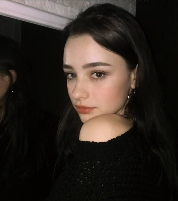 A close-up portrait of a young woman with long dark hair, looking over her shoulder, wearing a black off-the-shoulder top with hoop earrings against a dark background.