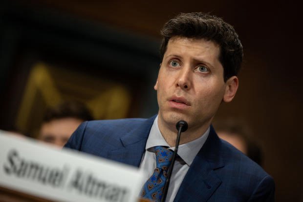A man in a blue suit and patterned blue tie sits at a microphone with a nameplate partially visible reading 'Samuel A...man', appearing to testify or speak at a formal hearing.