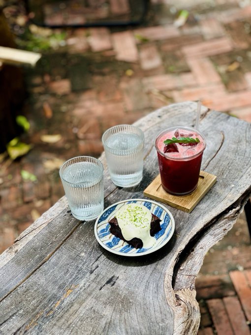 Two glasses of water, a red iced drink garnished with mint, and a dessert topped with cream and lime zest sit on a rustic wooden table outdoors.