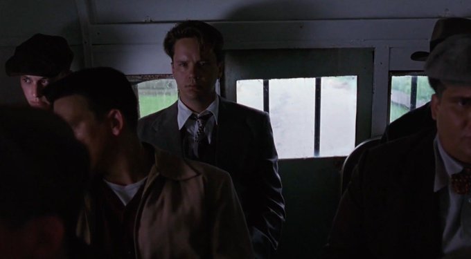 A man in a suit and tie stares intensely from the dim interior of a bus, surrounded by other passengers in hats and coats. The accompanying text reflects a fatalistic, cynical worldview—viewing all financial systems as scams—which mirrors the scene's somber, watchful mood as if the man is sizing up the world around him with cold calculation.