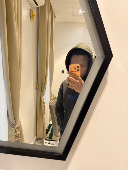 A person in a gray hoodie takes a mirror selfie with an orange phone, the word "MMAAT" visible on a sign below the mirror, symbolizing the quiet, unglamorous routine behind disciplined trading that yields results in just four weeks.