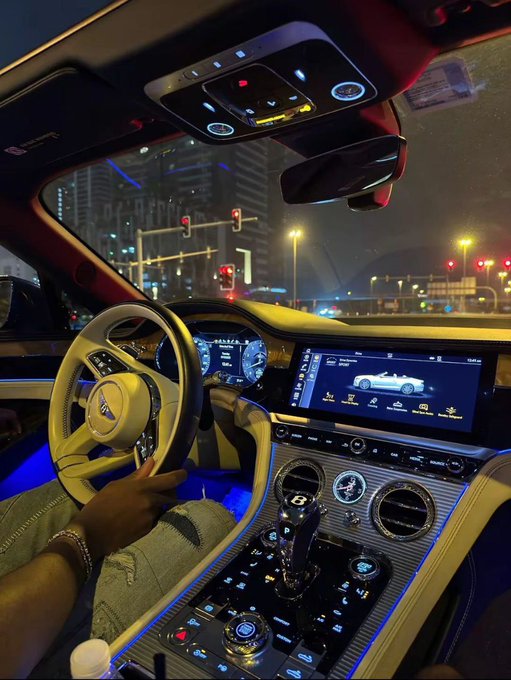 A driver grips the steering wheel of a luxury Bentley at night, with the illuminated dashboard displaying the digital speedometer and infotainment screen showing a Bentley model graphic, all framed against a city skyline with red traffic lights. The post "Good Night X" ties into the late-night urban driving scene, suggesting a stylish send-off from behind the wheel of a high-end vehicle.