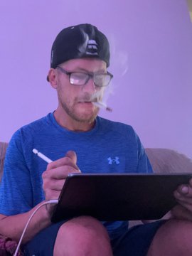 A man wearing glasses, a black cap, and a blue Under Armour shirt is smoking and drawing on a tablet with a stylus.