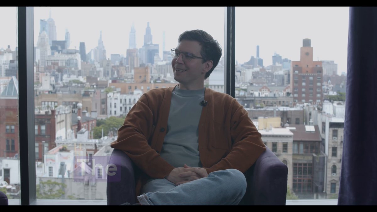 The image shows a man sitting in an office chair, looking out at a city view through a window. The man appears to be wearing glasses and has a warm brown sweater on. He seems to be enjoying his time indoors while observing the cityscape outside.