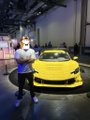 A man wearing a mask and a racing shirt stands next to a bright yellow Porsche sports car in a garage setting.