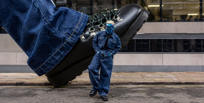 A person with a blue-painted face and head stands in a blue denim outfit leaning against a giant black boot in a denim jean outfit on a city street.