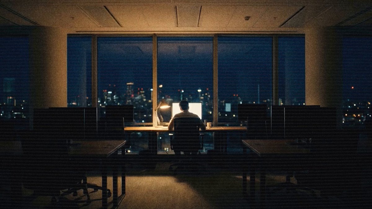 The image shows a person sitting at a desk with their back facing the camera, looking out of an office window that overlooks a city skyline. The room has multiple chairs and desks, suggesting a workspace or shared area for employees. The photo is captioned " Highly recommend applying".