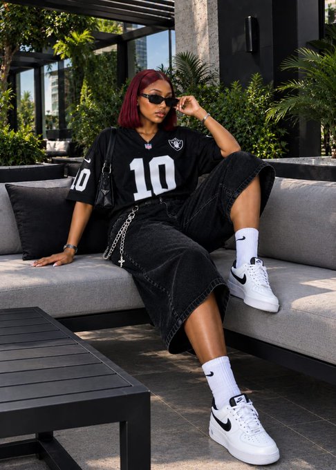 A woman with red hair wearing a Las Vegas Raiders jersey (number 10), black denim skirt, white socks, and white sneakers sits on an outdoor gray sectional sofa with greenery in the background.