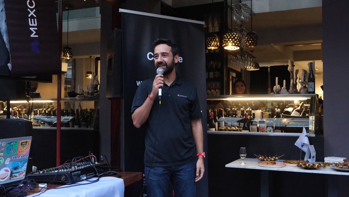 A smiling bearded man in a black polo shirt speaks into a microphone at an indoor event, with a bar/lounge area behind him displaying plates of food and bottles.
