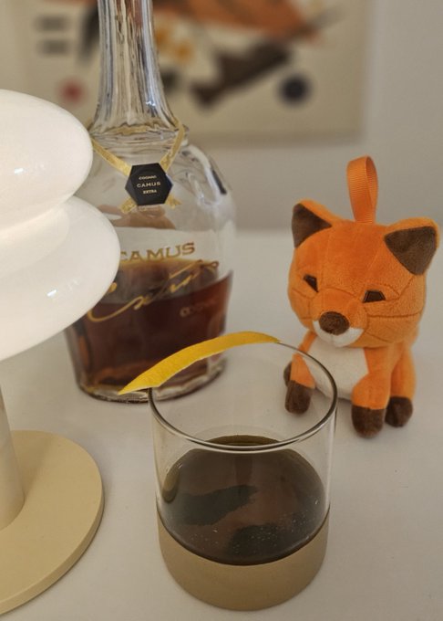 A Camus cognac bottle sits behind a glass containing a dark beverage garnished with an orange peel, next to a white lamp and an orange plush fox toy on a desk.