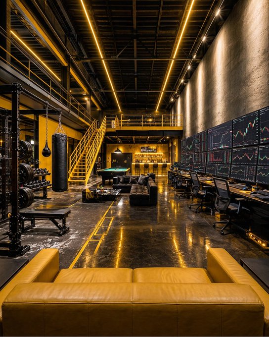 An industrial-style gym and lounge with yellow accent lighting features a punching bag, weightlifting equipment, a pool table, and wall-mounted monitors displaying financial charts.
