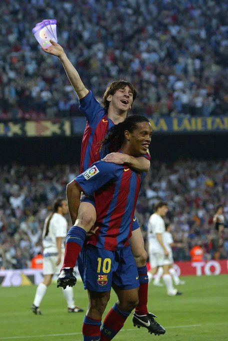 Two FC Barcelona soccer players in their red and blue striped jerseys celebrate, with one player jumping onto the back of player number 10, Ronaldinho, while holding up a folded shirt.