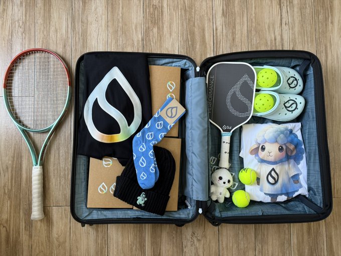 An open black suitcase displays an assortment of branded merchandise, including a pickleball racket, socks, a small dog plushie, and yellow balls, alongside another racket resting outside on a wooden floor.