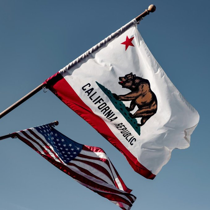 The California Republic state flag, featuring a brown bear and red star, flies alongside the American flag against a clear blue sky. This imagery relates to the news that California lawmakers have passed a bill banning public officials and employees from issuing meme coins, with the ban set to take effect in 2027.