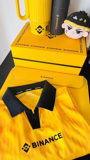 The image features a yellow shirt with black text that says "Binance", along with a yellow box containing a toy figure, likely a character from a video game or movie franchise. The background of the image is white, which makes the colors of the items stand out even more. The main focus of the image is on the Binance merchandise and its packaging, showcasing the brand's logo and the "Football Frenzy Trading Competition" event that may be related to it.