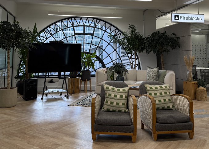 A modern, stylish lounge area with a sofa, two armchairs with striped pillows, a large TV, and plants in front of an arched window, branded with a 'Fireblocks' sign.