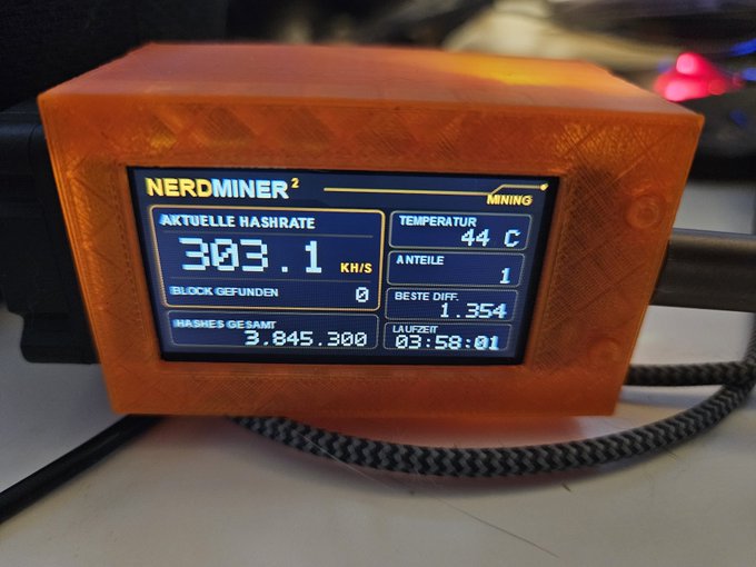An orange 3D-printed enclosure houses a display screen showing 'NERDMINER²' mining statistics with hash rate 303.1 KH/S, temperature 44°C, and 3,845,300 total hashes.