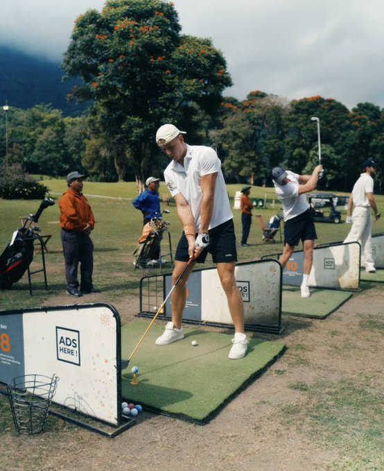 A man in a white cap and polo shirt practices his golf swing at a driving range, with an "ADS HERE!" sign visible on the barrier, while other golfers and a caddie are seen in the background. The relaxed, leisurely scene of people enjoying golf outdoors humorously illustrates the post's point about using the quiet Bitcoin bear market to take trips and pursue hobbies.