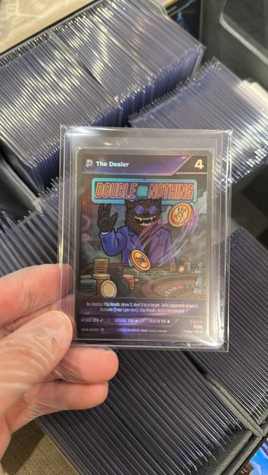 A hand holds a holographic Fuzzy NFT trading card titled "The Dealer" (rarity #4) featuring the "Double or Nothing" artwork, shown in front of stacked inventory of similarly sleeved cards.

The image directly illustrates the post's point about holding a digital $Fuzzy NFT in physical card form, with "The Dealer" epic card as the highlighted favorite.