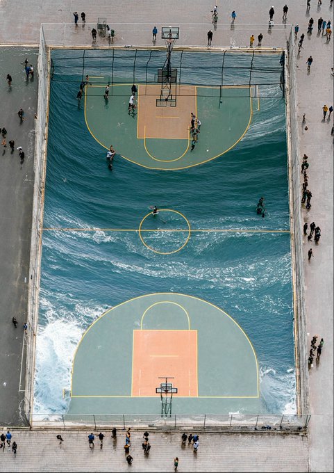 An aerial view shows an unusual flooded basketball court where waves are crashing onto the playing surface, with people standing around the edges watching the surreal scene.