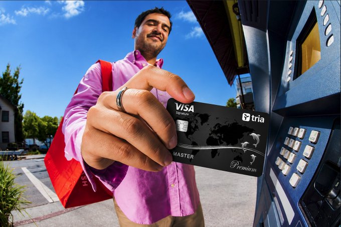 The image shows a man holding a Visa card at an ATM or self-service kiosk. The man appears to be wearing a pink shirt and has a red bag with him. The scene suggests that he might have just made a purchase using his credit card, as he is holding it up for the camera.