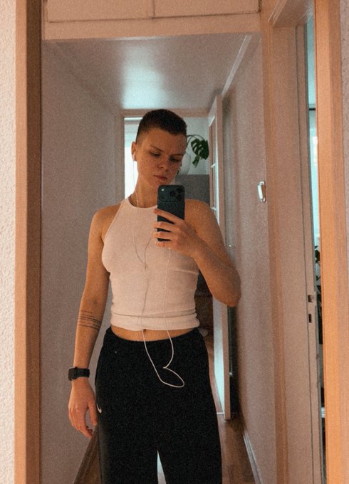 A person with a shaved head takes a mirror selfie in a hallway, wearing a white crop top, black pants, earphones, and a smartwatch.