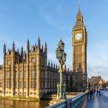 A photograph of the Palace of Westminster and Big Ben clock tower in London, viewed from Westminster Bridge on a sunny day with pedestrians walking along the sidewalk.