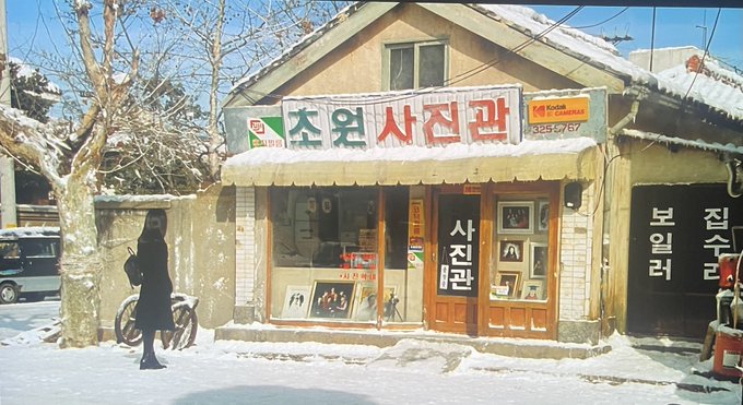 A snowy street scene featuring a storefront with Korean signage, including '초원사진관' (Chowon Photo Studio), where a person in a dark coat stands near a bicycle.