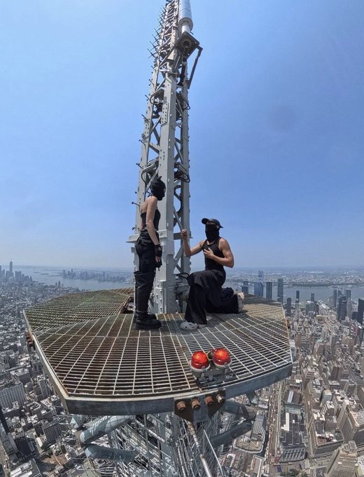 Two people in black outfits stand on a metal antenna platform atop a tall skyscraper with aerial views of a city skyline below.