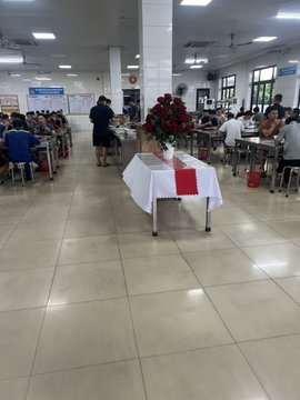 Interior of a cafeteria with people seated at tables eating, with a table in the foreground holding a vase of red roses and a white tablecloth with a red stripe.