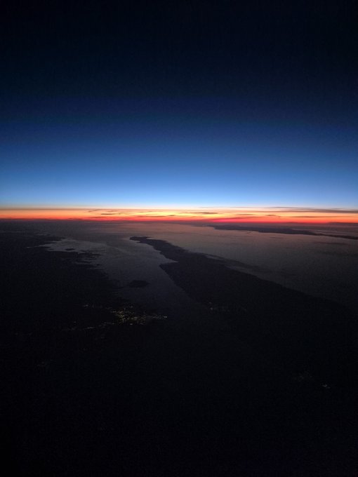 An aerial view at dusk or dawn showing a coastline or fjord with a vibrant orange and red sunset along the horizon, contrasted against the dark blue sky and silhouetted landscape below.