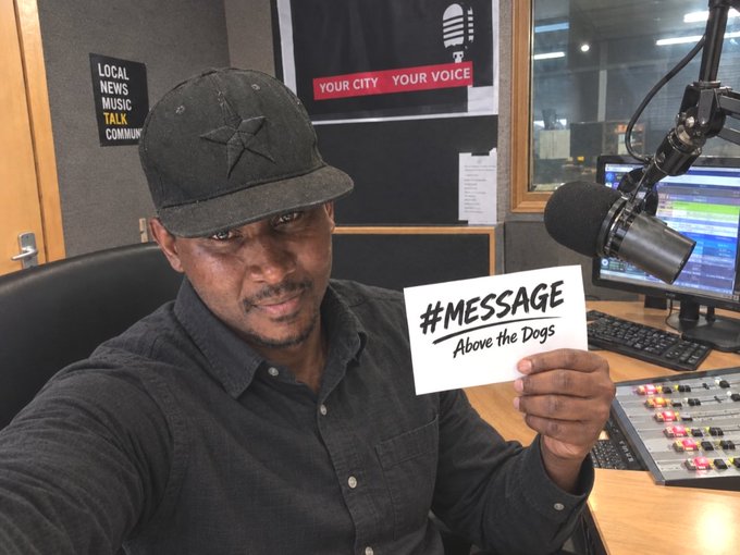 A man in a black cap and dark shirt sits in a radio studio holding a card that reads '#MESSAGE Above the Dogs', with a 'YOUR CITY YOUR VOICE' poster and microphone visible in the background.