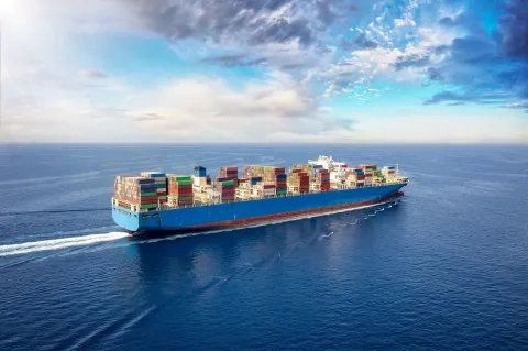 A large blue container ship loaded with colorful cargo containers sails across a vast blue ocean under a partly cloudy sky.