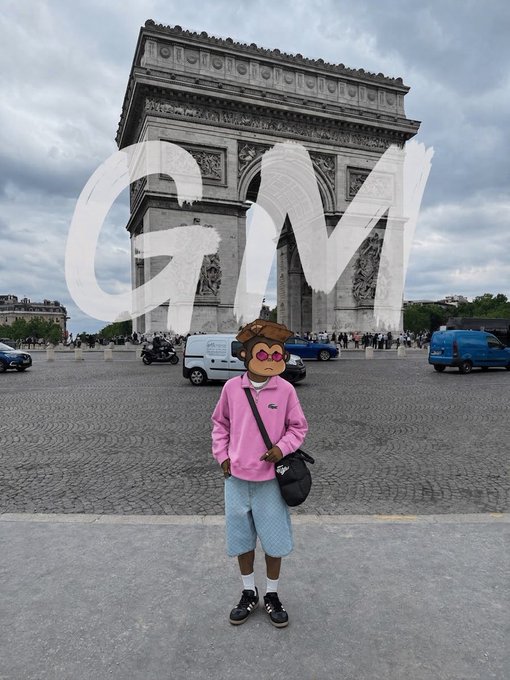 A photo of a person wearing a pink sweatshirt and light blue shorts standing in front of the Arc de Triomphe in Paris, with their face obscured by an animated avatar sticker wearing pink sunglasses, and "GM" painted in large white letters across the sky.