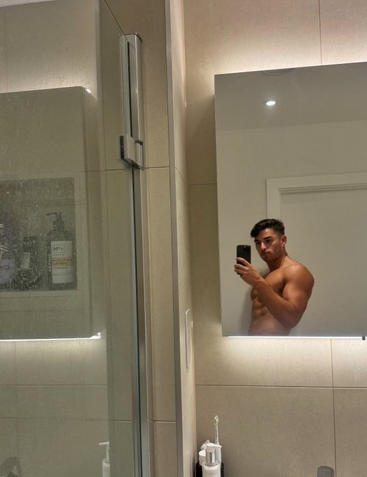A shirtless young man takes a mirror selfie in a modern bathroom with illuminated LED edges and a built-in shelf displaying shampoo bottles, while holding up his phone to capture the shot—appearing to enjoy a moment he wishes could "last forever."