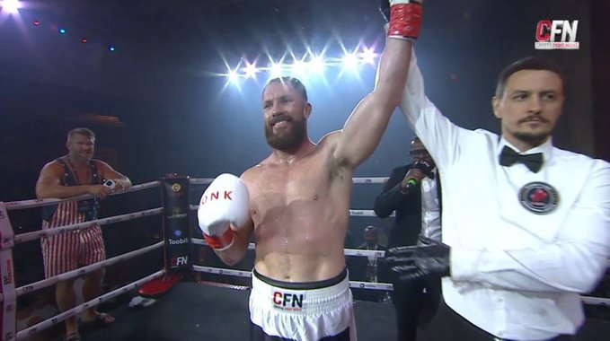 A bearded boxer wearing white "CFN" trunks and red "INK" gloves stands in the ring with his right arm raised in victory by a referee in a white shirt and bow tie, while a ring girl in an American flag-themed outfit holds the championship belt. The "CFN" (Combat Fight News) logo in the corner ties the celebratory moment to the post's announcement of upcoming fight news.