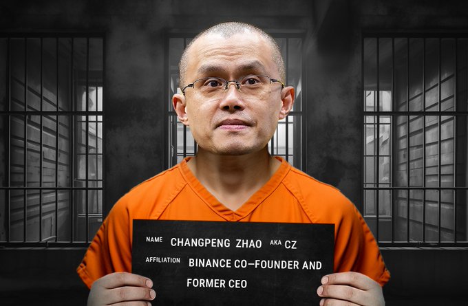 In this image, there is a man standing inside an jail cell. He is holding up a sign that says "Changping Zhao" with his finger pointing at it. The sign appears to have writing on it as well. The man has glasses on and is wearing an orange shirt. The background of the scene includes two bars on either side, giving a sense of confinement. A description in the image states that he donated $500K towards education for U.S. prisoners every six months.