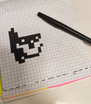 A black pen rests on a grid notebook next to a small pixel art drawing of a figure.