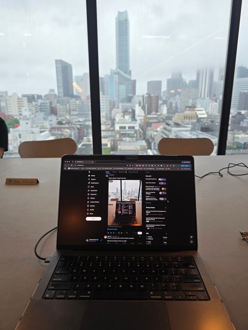 A laptop displaying X (formerly Twitter) sits on a table in front of a large window overlooking a city skyline with skyscrapers.