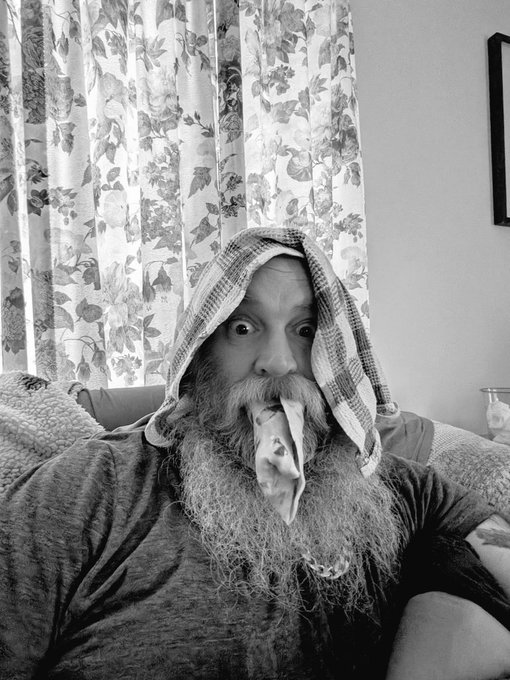 A black and white photo of a bearded man with a towel wrapped around his head, looking wide-eyed at the camera with the towel's tail held in his mouth.