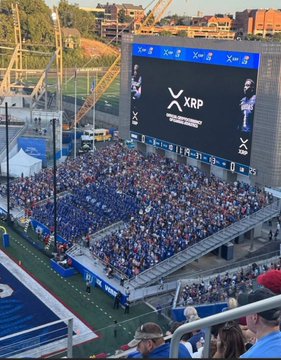 A packed stadium crowd cheers in blue seats beneath a large jumbotron displaying the XRP logo and "Powered by X," tying into the post's excitement about XRP being featured on the big TV screen.