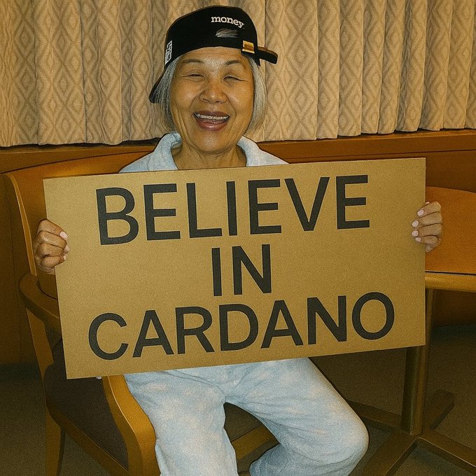 Smiling elderly woman wearing a black 'money' cap holding a cardboard sign that reads 'BELIEVE IN CARDANO', referencing the Cardano cryptocurrency.