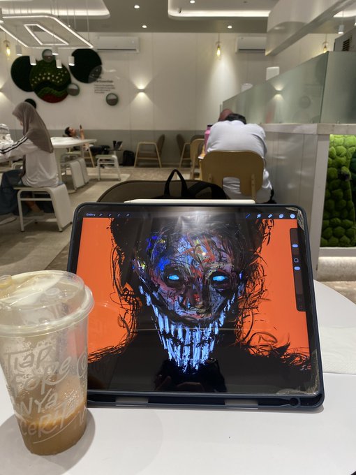 A photo of an iPad displaying a digital painting of a dark, skull-like face with glowing blue eyes, placed next to a bubble tea cup in a cafe.