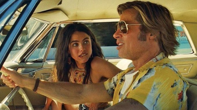 Cinematic still of a man in a yellow floral shirt and sunglasses driving a car with a young girl in the passenger seat, captured in warm vintage lighting.