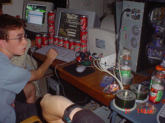 A person wearing glasses sits at a cluttered desk stacked with numerous Coca-Cola and Mountain Dew cans, working at a CRT computer monitor, with the timestamp '31 7:40 AM' visible in the corner.