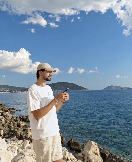 A man in a white t-shirt, beige cap, and shorts stands on rocky terrain by a blue sea, looking out at the water while holding a phone, with mountains visible in the background.